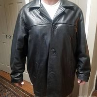 Front view of men's black leather coat worn by model, showing collar, button closure, and side pockets.