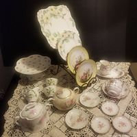 Wide view of the entire china collection on lace tablecloth showing plates, cups, cake stand, teapot, pitchers, and sugar bowls.