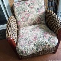 Front angle shot showing floral cushioned seat and backrest on rattan braided wicker chair with wooden armrests.