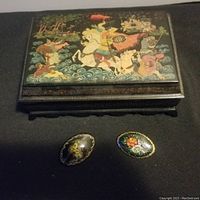 Top view of rectangular black paper mache lacquer box with painted historic battle scene and two oval floral brooches in front