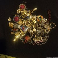 Pile of assorted vintage scrap jewellery on a black surface including rings, chains, brooches, and decorative pieces with mixed metals and faux stones.