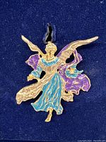 Close-up photo of the angel ornament showing the colored enamel robes in blue and purple and gilt metal wings posed in flight, mounted in a dark navy velvet lining inside the original box.
