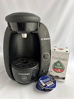 Front view of Bosch Tassimo coffee maker with drip tray and power button, accompanied by Nabob coffee pod box and two Maxwell House pods in front.