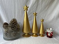 Photo showing three gold glitter Christmas trees with star toppers in descending size, a frosted glass bowl with pinecones inside, and a red bud vase with silver star detail.