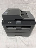 Front view of Brother MFC-L2740DW printer showing top scanner lid, control panel with small display, and paper output slot.