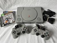 Photo showing the Sony PS1 console with power and open buttons, 2 wired controllers, power adapter, and stack of game cases including Tomb Raider and NFL GameDay 98.