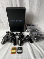 Full view of the PlayStation 2 console with two controllers, two memory cards, and cables included in the lot.