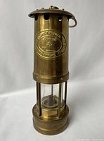 Full front view of the vintage brass Cambrian miners lamp with embossed nameplate and glass chimney visible.