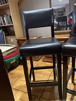 Front angled view of one black vinyl upholstered wooden bar stool showing the seat, backrest and legs.