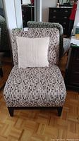 Front view of the patterned upholstered armless chair with beige decorative pillow on seat and dark wood legs. Shows the chair fabric pattern and pillow texture.