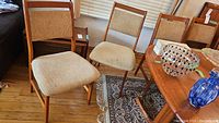 Four wooden chairs with beige fabric upholstery arranged around a table, showing overall lot.