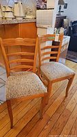 Two wooden dining chairs with ladder style backs and beige textured upholstery on seats, set on a hardwood floor by a kitchen counter
