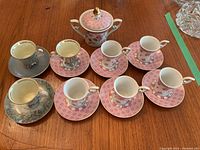 Full set of espresso cups, saucers, and sugar bowl arranged on wooden surface showing both pink floral and gray silver designs with gold trimming.