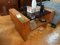 Wood coffee table with glass top, visible stains and wear on the wood sides, rectangular shape, lower wood shelf is visible with items stored underneath.