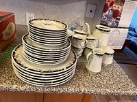 Full view of stacked plates, cups, teapot without lid, creamer and sugar bowl, all on countertop showing the pattern and amount of items
