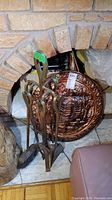 Fireplace tool set with poker, brush, shovel, standing on metal holder near fireplace, with round decorative copper plate behind it.