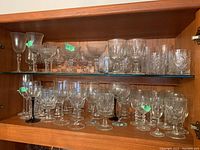 Glassware collection featuring various goblets, wine glasses, port glasses, and crystal whiskey glasses arranged on wooden shelves inside a cabinet.