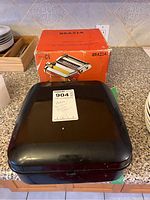 Closed black metal case and original orange box of Grazia pasta maker on a kitchen counter