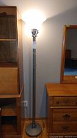 Full view of torchier style metal floor lamp with glass shade illuminated, showing overall height and setting beside wooden furniture.