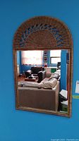 Full view of the wall mounted rectangular mirror with a semi-circular brown wicker frame, mounted on a blue wall reflecting living room furniture and decor.