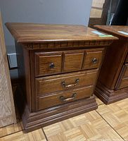 Wooden nightstand with two drawers, top drawer has two round metal knobs, bottom drawer has two metal handles. Decorative carved trim on top edge. Dark brown wood finish.