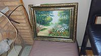 Full view of the framed painting displaying a garden path and flowers, resting against a wall and chair.