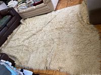Full view of the large off-white wool flokati style rug laid out on wooden floor between couches and other furniture.