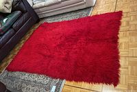 Full view of red wool flokati style rug on hardwood floor with other small rugs partially visible around it.