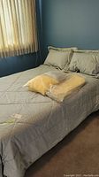 Bed set with light-colored bedding, pillows, and sealed plastic bag containing extra bedding item on top.