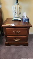 Wooden bedside table with two drawers, brass handles, and a decorative lamp on top.