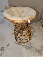 Front view of rattan papasan chair with beige cushion and rattan ottoman on concrete floor.