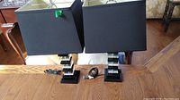 Two table lamps side by side on a wood table, showing clear view of black square lampshades and transparent acrylic bases stacked in blocks on black platforms. Power cords are visible coiled next to the lamps.