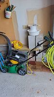 Green and black electric lawn mower with yellow extension cord wrapped around the handle, placed next to a garden hose and concrete blocks, indoor garage setting.