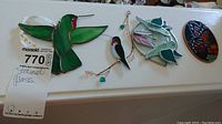 Four stained glass window hangers displayed on a white shelf including a green hummingbird, small bird, larger hummingbird with pink and green glass pieces, and a butterfly.