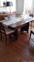 Dining table with glassware on top and chairs surrounding it, showing wear and fabric seat cushions.