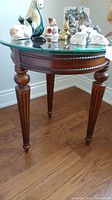 View of one wood and glass side table showing carved wooden legs and rope-like trim under glass top, placed on wooden floor.