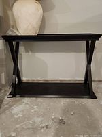 Front view of the dark wooden side table standing on a concrete floor, showing the tabletop and lower shelf connected by X-shaped side supports.