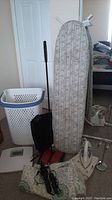 Photo showing ironing board with patterned cover, plastic laundry basket, white clothes iron, floor sweeper, bathroom scale, and electric back massager.