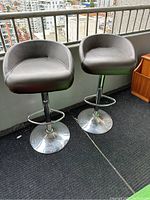 Set of two black faux leather bar stools with chrome bases and curved backs placed outdoors on balcony with city view.