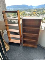 Two book shelves placed side-by-side on a balcony with mountain and city views in the background. One is a taller vintage wooden bookshelf with decorative side panels, the other a shorter pressboard bookshelf.