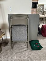 Folded card table and four folded chairs stacked against wall with bridge mat on floor