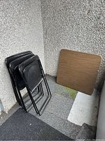 Four black folding chairs stacked near the wall next to folded wood-grain square card table.