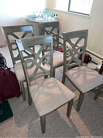 Four wooden dining chairs with beige fabric seats, decorative backs in geometric loop pattern, arranged closely together in a room.