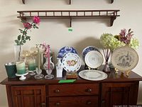 Wide view showing the assortment of plates, candles, artificial flowers in vases, and a wooden wall shelf above on a wooden sideboard.