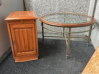 Image showing wooden cabinet/end table with drawer and door on left, and round glass coffee table with metal frame on right, against a grey textured wall and dark carpet.