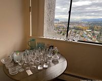 Photo of a wood round table by window showing collection of crystal glasses, tumblers, candlesticks, and dishes with city view background.