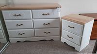Photo showing full view of the wood dresser and matching nightstand against a wall. The dresser features six drawers with brass handles, painted white with a natural wood top. The nightstand has three drawers matching the dresser style.