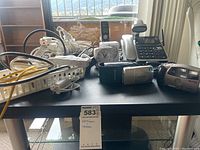 Photo of table top with multiple electronics including phones, extension cords, cameras, and small clock