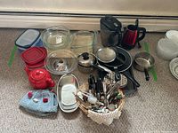 Wide shot of assortment of kitchen items on carpeted floor including glass bowls, pots, pans, utensils in basket, electric kettle and food storage containers
