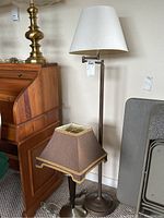 Photo of two lamps side by side: one tall metal floor lamp with white shade; one smaller metal table lamp with brown shade and gold trim.